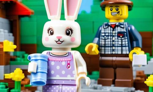 Il personaggio principale è una piccola coniglietta LEGO bianca dalle grandi orecchie morbide e occhi brillanti, timida ma rassicurata, che tiene un biglietto disegnato; indossa un vestitino lavanda a pois rosa e tiene una copertina blu ricamata e un peluche LEGO nell’altra zampa. Il padre coniglio LEGO, una figura alta col cappello a quadri e sorriso dolce ma preoccupato, è inginocchiato accanto a lei per abbracciarla; la madre è assente ma suggerita da una piccola valigia gialla aperta vicino alla porta con l’etichetta “Lilla è sempre amata”. L’ambiente è una tana LEGO luminosa e colorata con muri lavanda pastello, scaffali di piccoli libri illustrati, un angolo letto con coperta blu e tappeto floreale e una finestra rotonda sul prato; la scena è un momento intimo e dolce, con giochi e disegni appesi e luce dorata che attraversa la finestra. Lo stile visivo: colori vivaci, texture nette dei mattoncini, composizioni semplici ed espressioni esagerate e leggibili per trasmettere emozione.