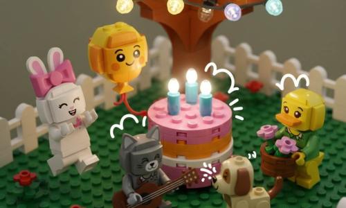 Sunny is a round yellow-and-orange brick balloon with a painted smiling face, floating slightly and reaching a ribbon-paw toward a pink-and-orange brick cake with three lit blue translucent candles and wisps of translucent white smoke; Rosie, a white-brick rabbit with a pink bow, is jumping and clapping left of the cake; Daisy, a yellow-and-green brick duck with textured feathers, holds a small flower basket right of the cake; Sam, a gray-brick cat with a brown mini-brick guitar, sits playing happily; Popcorn, a brown-and-cream brick puppy with a wagging tail, looks at the cake as if he sneezed and blew out the candles; the garden set has a textured green lawn, a brown-and-green brick tree with multicolored translucent string lights, a few brick flowers and a low white fence, creating a warm, lively celebration seen from a slight bird’s-eye angle.