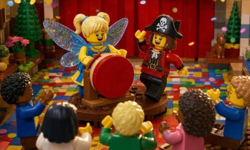 Due bambine di circa 7 anni: una bionda con due codini in costume da farfalla blu e giallo con ali traslucide che sorride al pubblico mentre tiene un grande tamburo rosso davanti a sé al centro-sinistra del piccolo palco, l'altra castana con taglio a caschetto in costume da pirata rosso e nero con un ampio cappello nero che danza a destra del palco agitando la mano e ridendo verso l'amica. La scena è una piccola sala per feste costruita in mattoncini LEGO con pavimento a scacchi colorato, ghirlande trasparenti multicolori, un piccolo palco in mattoncini al centro, panchine con minifigure che applaudono, un grande tendone rosso sullo sfondo e coriandoli a pezzi rotondi sparsi; le bambine fanno un numero di carnevale: la tamburellista suona dolcemente mentre la pirata invita il pubblico, luce calda e movimento suggerito dai pezzi inclinati e dai coriandoli sospesi.