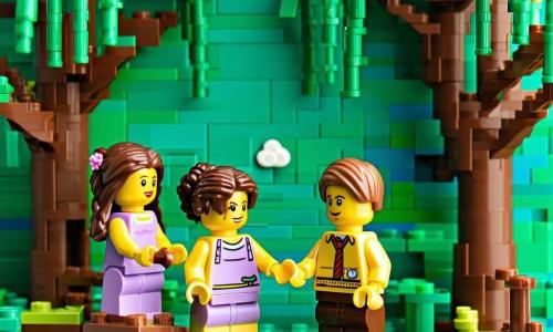 Quattro bambine di 5 anni sedute sui grandi e rotondi rizomi di un salice piangente in mattoncini verde scuro e marrone, da sinistra: Lila capelli castani in due trecce con vestitino viola che tiene la mano di Marta e guarda il cielo; Marta capelli neri ricci con vestito giallo che tiene una lente rossa giocattolo mentre osserva un’formica di mattoncini; Sofia capelli biondi in coda con vestito blu, mani sulle ginocchia, che ascolta il vento; Emma capelli rossi corti con vestito verde che tiene una piccola mela di mattoncini accanto a un riccio LEGO con un’altra mela. Scena al crepuscolo in un parco con uno stagno a destra che riflette il cielo rosato, un melo sullo sfondo sinistro carico di mele rosso‑arancioni, prato a lastre verdi con qualche fiore giallo e blu, vento visibile in sottili traiettorie curve che sollevano capelli e foglie, luce morbida e rosata, luna all’orizzonte, stile LEGO dettagliato, angolazione leggermente dall’alto con messa a fuoco nitida sui personaggi e sfondo leggermente sfocato.