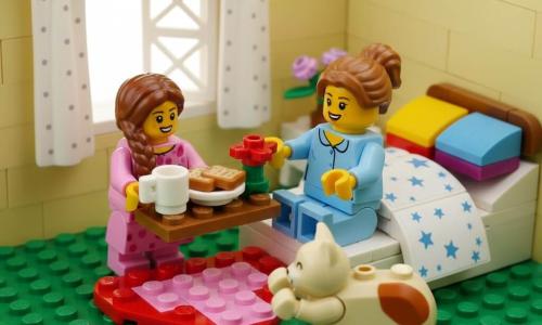 Menina de 6 anos alegre e travessa, cabelos castanhos longos em tranças, olhos brilhantes, vestido rosa com bolinhas brancas, leva num tabuleiro um café da manhã com xícara, torrada e uma flor vermelha; mãe de cerca de 32 anos, emocionada e sorridente, cabelo castanho preso em coque, pijama azul claro, sentada na cama com as mãos estendidas, olhar amoroso; gato redondo e fofo chamado Bolinha, creme com manchas ruivas, perto da mãe com as patas esticadas; quarto iluminado de manhã com paredes amarelo pálido, janela aberta com cortina branca de bolinhas, vaso com pequenas flores no criado-mudo, cama com edredom branco de estrelas, almofadas coloridas e tapete em forma de coração; cena mostra a menina andando na ponta dos pés para surpreender a mãe; estilo diorama LEGO nítido, peças visíveis, cores vivas saturadas, texturas lisas, iluminação suave e composição centrada.