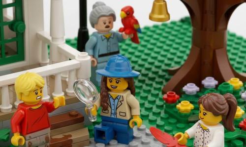 A young girl detective (about 10) kneels on a small brick porch examining muddy footprints with a translucent magnifying glass in her right hand; she has brown braided hair, a wide blue hat and a beige jacket. A blond boy, Oscar (about 8), stands left, out of breath and worried, pointing toward the garden, wearing a red t‑shirt and muddy sneakers. A brown‑haired girl, Lily (about 9), crouches by a flowerbed with a small red feather stuck to her shoe, smiling shyly in a polka‑dot dress and knee pads. Mr. Benson (about 60) is in the background in his garden with a red parrot on his shoulder, looking surprised. Mrs. Willow (about 70) stands on the porch with hands clasped, in a floral dress and shawl, relieved as she waits for the missing golden bell. The setting is a residential LEGO brick street: small porch with white railing, slightly muddy steps, green bushes, an old oak with a small LEGO squirrel holding a golden bell on a branch, a vintage lamppost and a paved path. Main scene: the detective studies muddy prints and the red feather on the porch while children and neighbors gather around; bright clear morning, vivid contrasting colors, sharp details, playful warm style, composition centered on the magnifying glass and the golden bell in the tree.